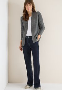 Šedý žebrovaný kardigan přes bílou košili, doplněný tmavě modrými rozšířenými džínami a světlými teniskami. Hladká textura s volným střihem.