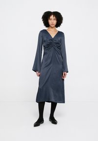 Rochie din satin bleumarin cu mâneci lungi, decolteu în V și detaliu adunat în centru. Textură netedă, lungime medie, asortată cu colanți negri și balerini.