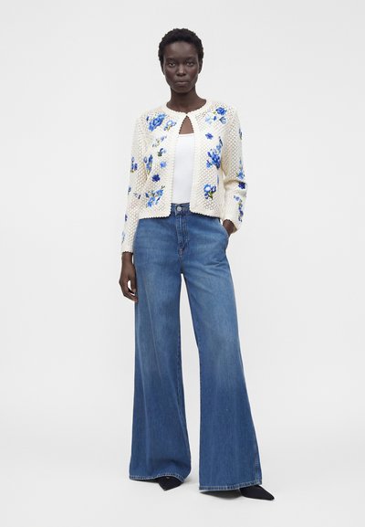 Kvinna som bär vida blå jeans, vit topp och krämfärgad stickad kofta med blå blommig brodyr, stående med en hand i fickan.