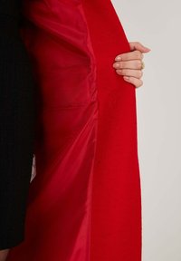 Red wool coat with a satin interior. The coat is unbuttoned, revealing the smooth lining and textured outer fabric. Accented with a black garment.