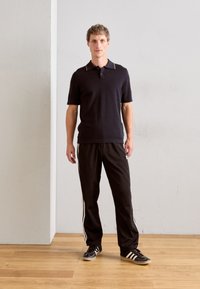 Polo noir avec un col contrasté, associé à un pantalon de jogging noir avec des rayures blanches sur les côtés. Le modèle porte des chaussures de sport noires.