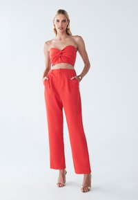 Červený strapless crop top s prekrúteným predným detailom a vysokými širokými nohavicami, v kombinácii s kovovými sandálmi na opätku s otvorenou špičkou. Hladká textúra látky.