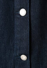 Camisa de mezclilla azul oscuro con botones plateados en la tapeta, que presenta una textura clásica y detalles de costura visibles cerca de los botones.