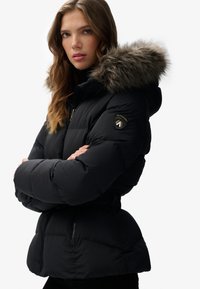 Giubbotto imbottito nero con cappuccio foderato in pelliccia, vita elastica e tasche laterali con zip; presenta una patch logo sul maniche.