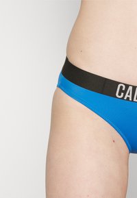 Slip bikini blu con una fascia elastica nera caratterizzata da scritte bianche. Tessuto liscio, design aderente e taglio a vita media.