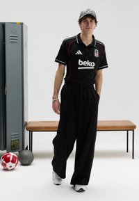 Juoda futbolo apranga su raudonomis detalėmis, derinama su plačiomis juodomis kelnėmis. Baltos sportbačiai, pilka kepurė. Fone matyti suoliukas ir sporto kamuoliai.