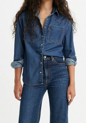 Femme portant une chemise en jean bleu foncé avec manches retroussées et un jean taille haute assorti, debout devant un fond clair uni.