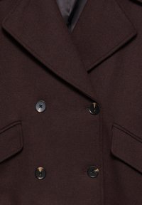 Valkoinen villakangasviitta, jossa on suuria mustia nappeja, terävä kaulus ja sivutaskut, jossa on kalastajapohjainen rakenne.