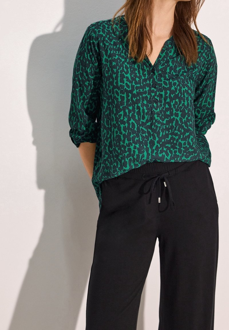 Femme portant un chemisier boutonné vert et noir à motifs avec des manches retroussées et un pantalon noir à cordon, debout contre un mur uni.