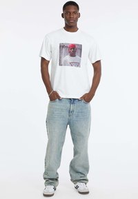 Biała bawełniana koszulka z dużym graficznym nadrukiem z przodu, zestawiona z luźnymi niebieskimi dżinsami i białymi trampkami w czarne paski.