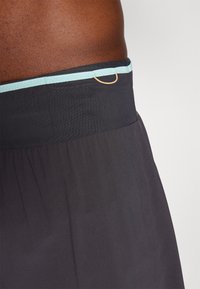 Bañadores negros con una cinturilla suave y elástica. Presentan un acento azul claro en la parte superior y un detalle en forma de anillo dorado en la parte frontal.