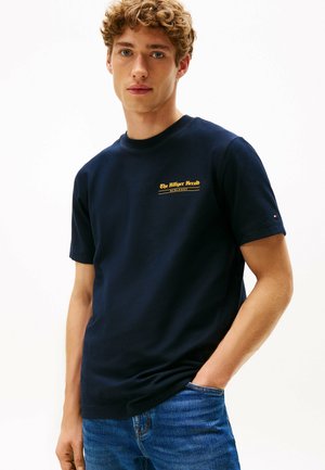 Ung mand med krøllet hår iført en navy blå T-shirt med gul tekst på brystet og blå jeans, stående med den ene hånd i lommen.