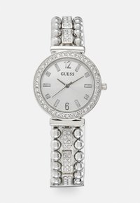 Montre-bracelet en argent avec un cadran rond, des chiffres argentés et des embellissements en cristal. Le bracelet présente des segments alternés de formes rondes et rectangulaires.