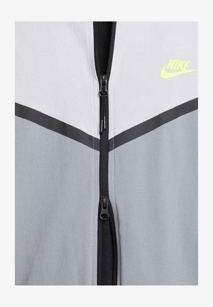 Chaqueta con cremallera de Nike con paneles en gris y blanco, que presenta una cremallera y costuras en negro. Logotipo en amarillo neón en la parte superior derecha. Textura suave.