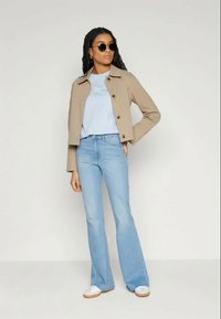 Beige cropped jakke med rund krage og sorte knapper, lyseblå t-skjorte, vide lyseblå jeans og hvite joggesko med brune såler.