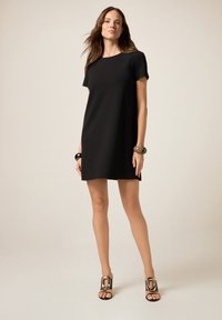 Schwarzes kurzes Kleid mit geradem Schnitt und rundem Ausschnitt. Das Model trägt ein voluminöses Armband und High Heels. Schlichte, glatte Textur.