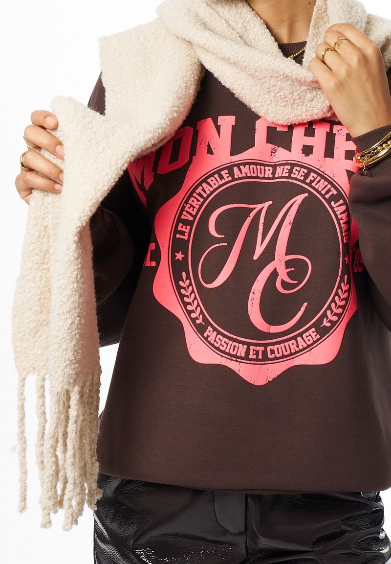 Sweatshirt marron avec impression graphique rose, associé à une écharpe blanche duveteuse affichant des bords à franges, porté par-dessus un pantalon noir brillant.