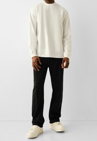 Man med vit långärmad skjorta, svarta byxor och off-white sneakers som står mot en enkel grå bakgrund.