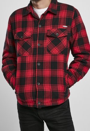 Uomo che indossa una camicia a quadri rossa e nera con tasche a patta, in piedi con le mani nelle tasche davanti a uno sfondo semplice.