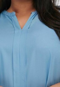 Světle modrá blůza s krátkým rukávem a výstřihem do V, s jemným nabráním v přední části. Vyrobena z hladkého, měkkého materiálu.