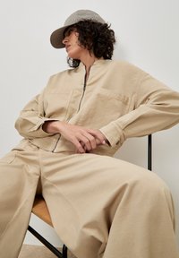 Persoon in een beige zip-up jacket, bijpassende wijde broek en een geruite pet, zittend met de handen in elkaar geklapt en naar de zijkant kijkend.