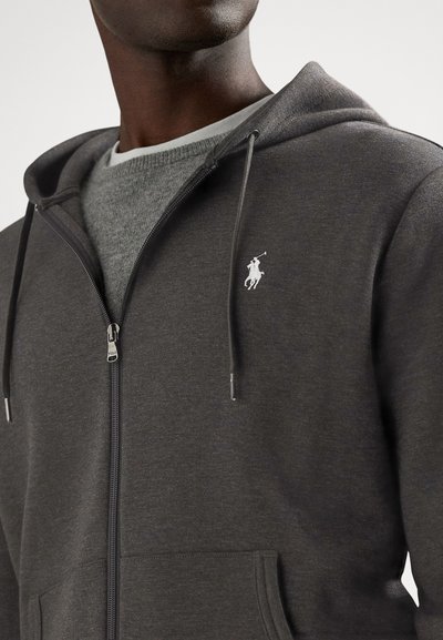 Γκρι φούτερ με φερμουάρ κατασκευασμένο από απαλό ύφασμα, με λευκό λογότυπο στο αριστερό στήθος, ρυθμιζόμενα κορδόνια και μπροστινές τσέπες.