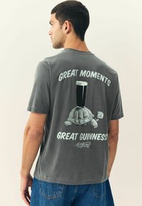Graue Baumwoll-T-Shirt mit einem Schildkrötenmotiv, auf dem ein Bierglas steht, und dem Text "GROßE MOMENTE, GROßES GUINNESS" in Weiß.