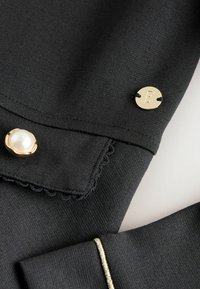 Melnā auduma ar viļņotām malām, ar zelta akcentiem, tostarp apaļš pogas ar 'B' un pērlēm līdzīga poga, demonstrējot tā gludo tekstūru.