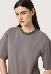 Maglietta grigia a maniche corte con colletto testurizzato e decorato con accenti metallici. Tessuto morbido e liscio con una vestibilità rilassata.
