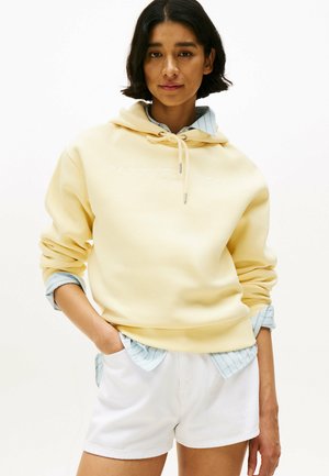 Een gele hoodie met een trekkoord en geborduurd logo, gelaagd over een lichtblauw gestreept shirt, gecombineerd met witte shorts.