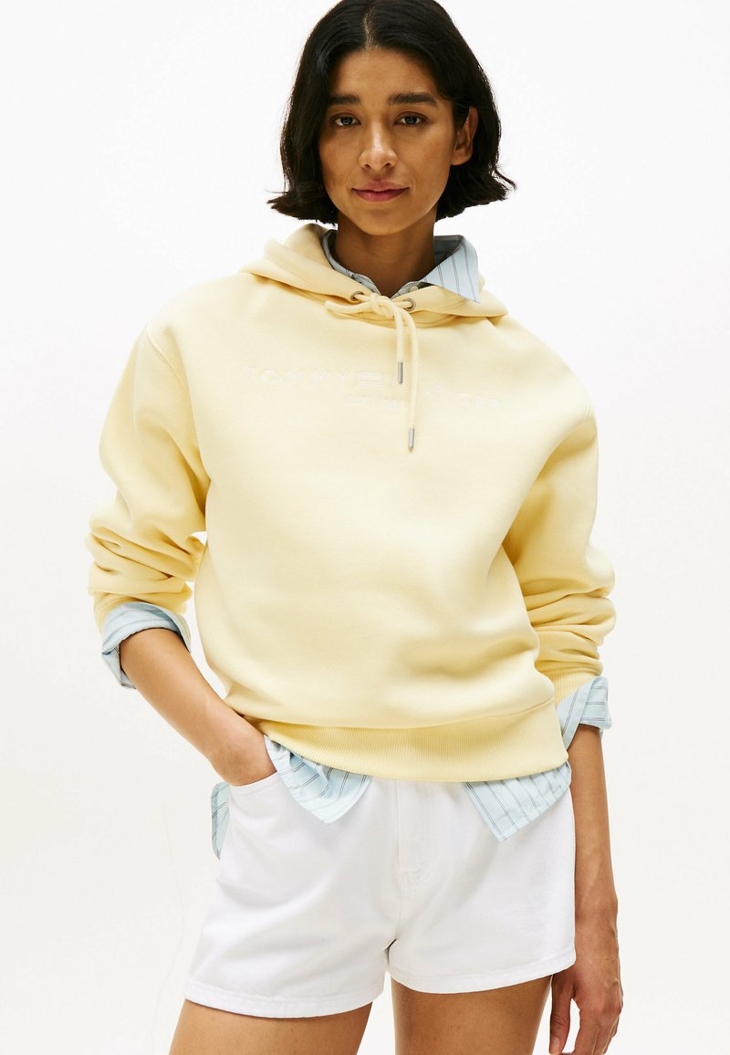 Felpa gialla con cappuccio e logo ricamato, indossata sopra una camicia a righe blu chiaro, abbinata a pantaloni corti bianchi.
