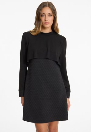 Frau trägt ein schwarzes, langärmliges Kleid mit gesteppter Rockpartie und einem überlappenden, mehrlagigen Crop-Top, steht vor weißem Hintergrund.