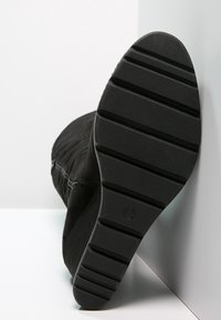 Bota de ante negro con suela de goma texturizada que presenta ranuras horizontales. Detalle de cremallera a lo largo del costado y una forma ligeramente curvada.