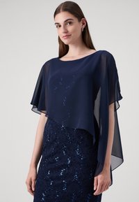 Marinblå chiffon cape topp med ett flödande design, prydd med subtila paljetter. Matchad med en marinblå spetskjol dekorerad med paljetter för textur.