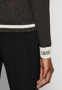 Haut noir scintillant à manches longues avec des rayures blanches accentuées au niveau du col et des poignets. Les poignets présentent le logo "LIU-JO" en noir.