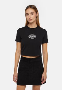 Camiseta corta negra de algodón, con un logo de globo bordado en blanco. El modelo la combina con una falda negra de pana.
