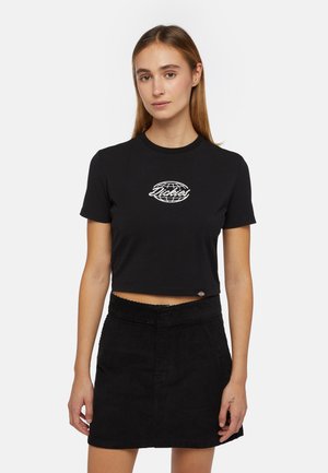 Zwarte cropped t-shirt van katoen, met een witte geborduurde wereldbol logo. Het model combineert het met een zwarte corduroy rok.
