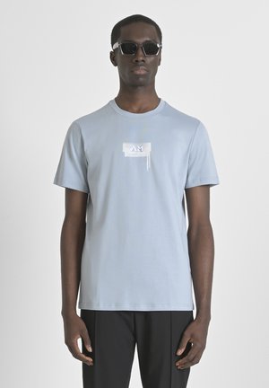 Camiseta de algodón de color azul claro con cuello redondo, mangas cortas y un acento de logo blanco en el pecho. El modelo lleva gafas de sol negras.