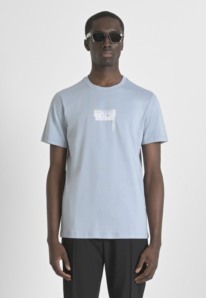 Camiseta de algodón de color azul claro con cuello redondo, mangas cortas y un acento de logo blanco en el pecho. El modelo lleva gafas de sol negras.