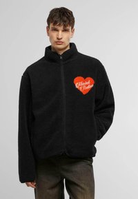 Black fleece jacket with a high collar, zip closure, and orange heart design with "Market Studios" lettering on the left chest.