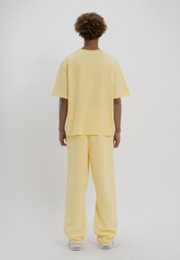 Une personne aux cheveux bouclés se tient de dos, portant un pantalon jaune ample, une chemise jaune surdimensionnée à manches courtes et des baskets blanches.