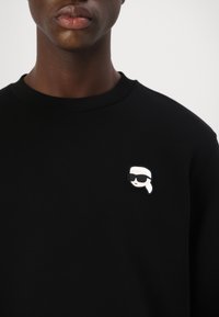 Črn pulover s okroglim izrezom, ki na levem delu prsi nosi majhno, barvito zaponko osebe v sončnih očalih.