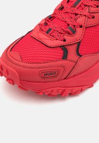Chaussure de sport rouge avec une tige en maille, des accents en caoutchouc et une semelle texturée dotée de crampons proéminents assurant une excellente traction. Logo sur le côté.