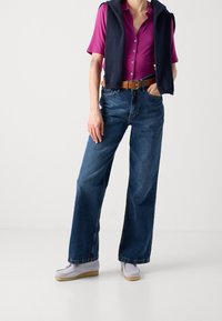 Casual outfit met een fuchsia shirt met korte mouwen, donkerblauwe wijde jeans en lichtgrijze instap sneakers met een tan kleurige zool. Marineblauwe trui over de schouder gegooid.