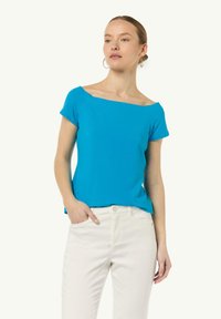 Krótki rękaw, bluzka typu off-the-shoulder w jasnym turkusowym kolorze, o dopasowanym kroju i miękkiej fakturze; zestawiona z białymi spodniami.