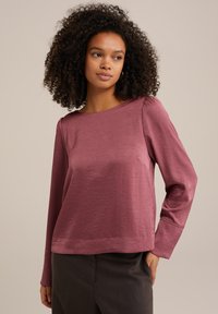 Halenka s dlouhými rukávy z jemné zmačkané látky v měkkém mauve odstínu. Má široký výstřih a jemné sklady na ramenech. Doplňuje ji tmavé kalhoty.
