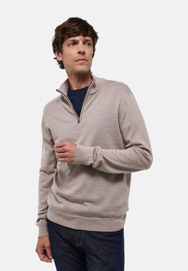 Pull à fermeture éclair quart beige clair avec des manches longues, des poignets et un ourlet côtelés ; porté sur un t-shirt bleu marine, texture lisse, design décontracté.