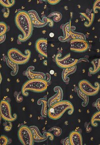 Camisa negra con un colorido patrón de paisleys en amarillo, verde y rojo, con botones blancos y un único bolsillo en el pecho.