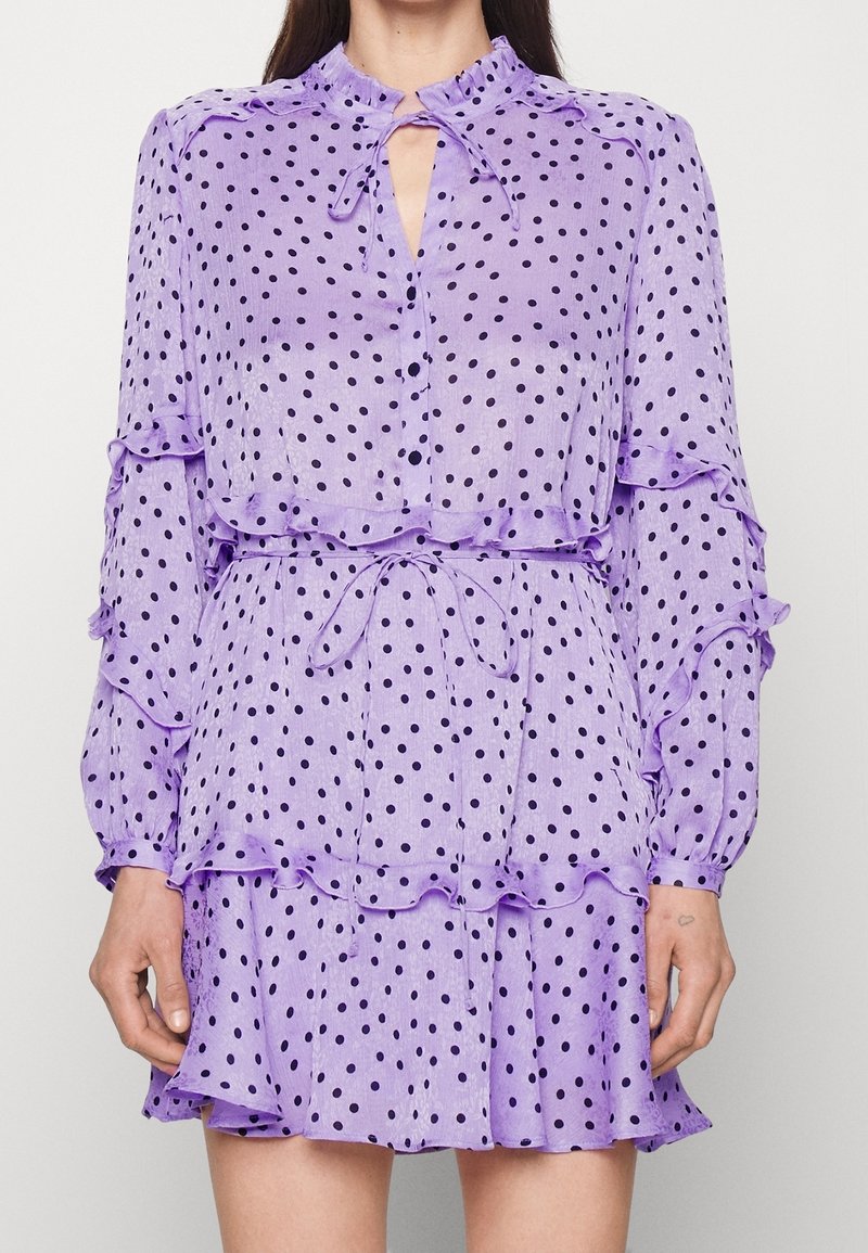 Robe à pois violets avec détails à volants et col à nouer. Confectionnée en tissu léger, avec des manches longues et une jupe évasée.