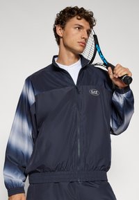 Navy windbreaker jacket with a gradient blue-gray pattern on sleeves, full zip closure, elastic cuffs, and EA7 logo on chest.